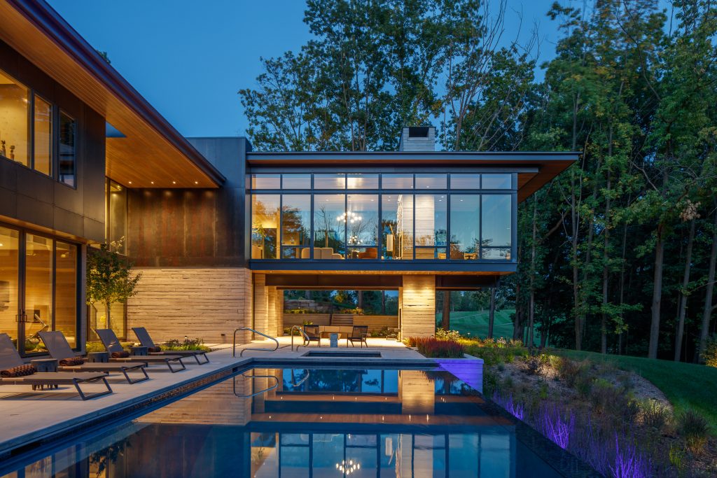 Luxury modern home designed by Lucid Architecture with large glass windows and an outdoor pool, surrounded by trees, photographed at dusk.