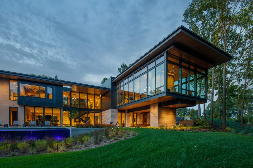 A luxury, modern two-story home designed by Lucid Architecture with large glass windows, illuminated from within, stands adjacent to a landscaped yard and pool at twilight.